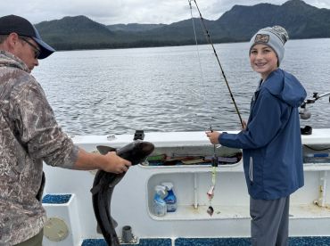A young angler smiles while holding a fishing rod, as an adult prepares to release a caught shark on a scenic fishing trip.