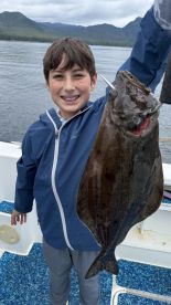 A young angler proudly displaying a Halibut while fishing on a boat in a scenic coastal environment.