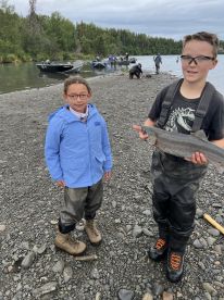 Two young anglers proudly displaying a freshly caught fish on a rocky riverbank, surrounded by boats and a lush green backdrop.