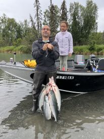 A proud angler displays a catch of multiple salmon while standing in shallow water next to a young girl, with a fishing boat in the background.
