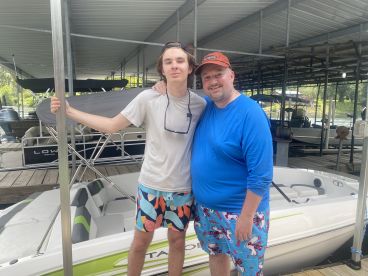 A father and son pose together in colorful swimwear next to their boat at a marina, showcasing a fun day on the water.