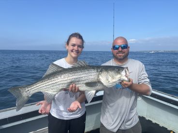 A young angler and an adult proudly displaying a large Striped Bass while fishing on a boat in open waters.