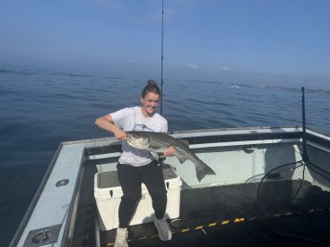 A young woman proudly displaying a large Striped Bass while fishing on a boat in calm waters.
