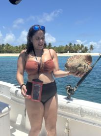 A woman holding a large shell while on a boat, with a tropical beach in the background.