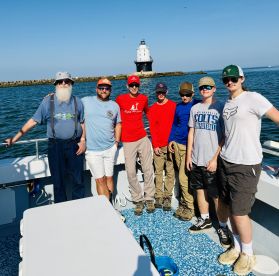 A group of anglers posing on the deck of a fishing boat, with a lighthouse visible in the background, enjoying a sunny day on the water.