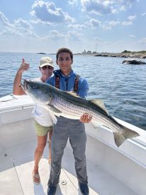 A young angler proudly displays a large Striped Bass while accompanied by an adult on a fishing boat, showcasing a successful day on the water.