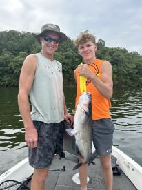 A young angler and an adult proudly display a large catfish while fishing on a lake, showcasing a successful day on the water.
