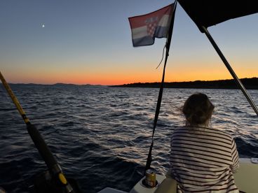A serene sunset view from the interior of a boat, showcasing a person enjoying the tranquil waters while a Croatian flag flutters in the breeze.