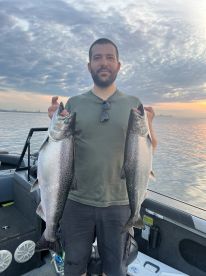 Angler proudly displaying two large Salmon while fishing on a boat during sunset.