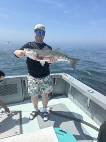 Angler proudly displaying a large Striped Bass while fishing on a boat in clear waters.