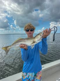 A young angler proudly displaying a large Speckled Trout while fishing on a boat in a scenic coastal environment.