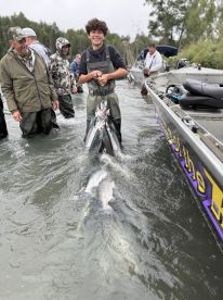 A young angler joyfully displaying a catch of salmon while standing in the water, surrounded by fellow fishermen during a group fishing trip.