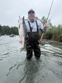 Angler standing in the water proudly displaying a large Salmon caught during a fishing trip in a scenic river setting.