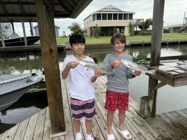 Two young anglers proudly displaying their catches of Speckled Trout at a dockside location, showcasing a fun day of fishing.