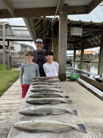 A group of two young anglers and an adult proudly posing with a neatly arranged catch of Speckled Trout on a cleaning table at a fishing dock.