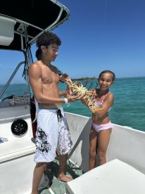 Two young anglers proudly displaying a large lobster aboard a boat in clear blue waters, showcasing a fun family fishing experience.