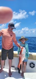 A young angler and an adult proudly displaying a large Red Snapper aboard a fishing boat in clear blue waters.