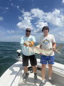 Two anglers proudly displaying a large shark on a boat during a fishing trip, with clear blue skies and calm waters in the background.