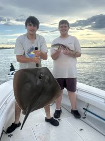Two young anglers proudly displaying a large stingray and a smaller fish while fishing on a boat in a scenic waterway.