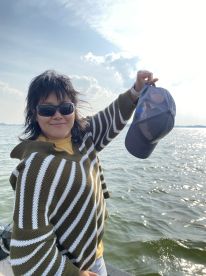 A person wearing sunglasses and a striped sweater is holding a cap while standing on a boat, with water and clouds in the background.
