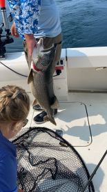 A young angler is focused on a large Lake Trout being held by an adult on a fishing boat, showcasing a successful catch during a summer outing.