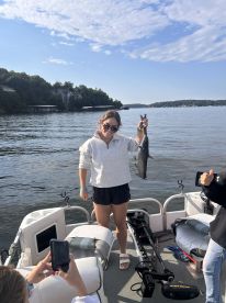 A young woman proudly displays her catch, a large fish, while on a boat in a scenic lake setting.