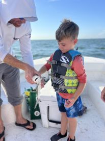 A young boy in a life jacket excitedly holds a small shark while an adult assists him on a boat, showcasing a memorable fishing experience.