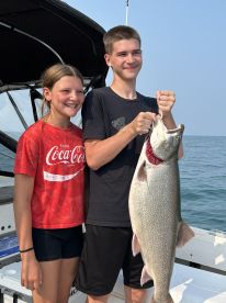 Two young anglers proudly displaying a large Lake Trout while fishing on a boat.