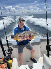 An angler proudly displaying a large Grouper while aboard a fishing boat, with a scenic ocean backdrop.