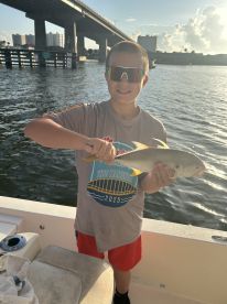 A young angler proudly displaying a fish while fishing from a boat, with a scenic backdrop of a bridge and waterfront buildings.