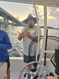 An angler proudly displaying a Yellowtail Snapper while fishing on a boat, with a scenic backdrop of a bridge and sunset.