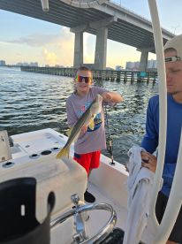 A young angler proudly displaying a Snook caught while fishing from a boat under a bridge, with a scenic waterfront backdrop.