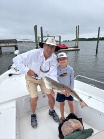 A proud grandfather and his grandson display a large Redfish on a boat after a successful fishing trip, showcasing a family bonding moment.