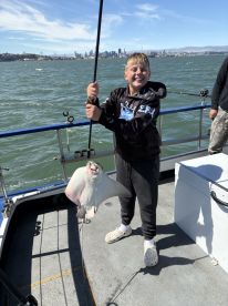 A young angler joyfully holding a caught stingray while fishing on a boat in a scenic bay area, with a city skyline in the background.