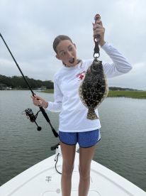A young angler excitedly holds up a Flounder while fishing from a boat in a calm waterway.