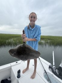 A young angler proudly displaying a large stingray while standing on the deck of a fishing boat in a grassy coastal area.
