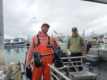 Two crew members smiling on the deck of a fishing boat, showcasing the interior setup with a view of the marina in the background.