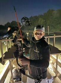 A young angler preparing to fish at night with a bow on a boat, showcasing the excitement of bowfishing in a serene water environment.