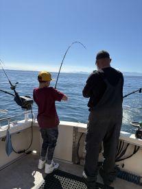 A young angler and an adult are engaged in fishing, both reeling in their lines on a sunny day, showcasing a vibrant fishing experience on the water.