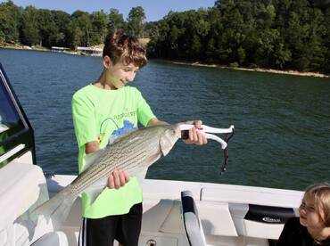 A young angler joyfully displaying a striped bass while fishing on a lake, with another child observing from the boat.
