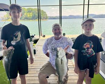 A proud moment at the dock with two young anglers holding their catches, a striped bass and another fish, after a successful day of fishing on a scenic lake.