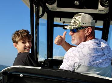 A young boy and an adult are enjoying their time on a boat, with the adult giving a thumbs up while navigating the waters.