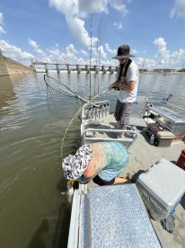 Two anglers actively fishing from a boat, with one preparing to land a catch while the other reels in. The scene captures the excitement of fishing in a river environment.
