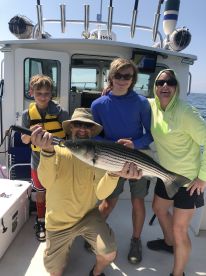 A family proudly displays a large Striped Bass aboard a fishing charter, enjoying a sunny day on the water.