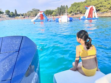 A young girl in a yellow swimsuit sitting on the edge of a boat, overlooking a vibrant water park scene with inflatable structures in the background.