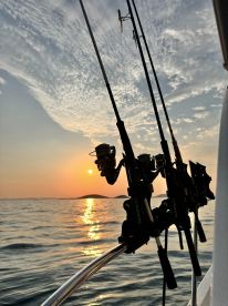 A serene sunset view from the side of a fishing boat, showcasing fishing rods against a backdrop of calm waters and distant islands.