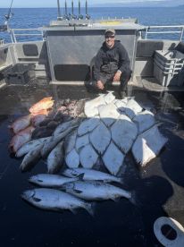 A smiling angler poses with a diverse catch including Halibut and Salmon on a fishing boat in open waters.