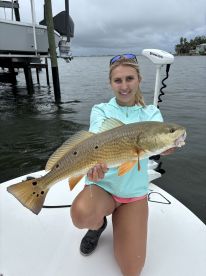 An angler proudly displaying a large Redfish while kneeling on the deck of a boat in a coastal waterway.
