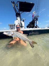 An angler proudly displaying a large Snook while wading in shallow waters, with a fishing boat in the background.