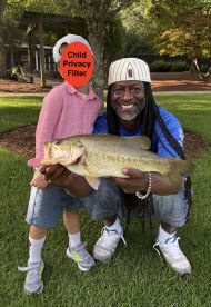 A proud angler holding a large Largemouth Bass while posing with a child in a grassy area, showcasing a successful fishing outing.